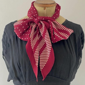 Puede incluir: Una bufanda burdeos con lunares blancos y un patr&oacute;n de rayas punteadas est&aacute; atada alrededor del cuello de un maniqu&iacute;. La bufanda est&aacute; hecha de una tela suave y fluida y est&aacute; anudada en un lazo. El fondo es un vestido gris oscuro.