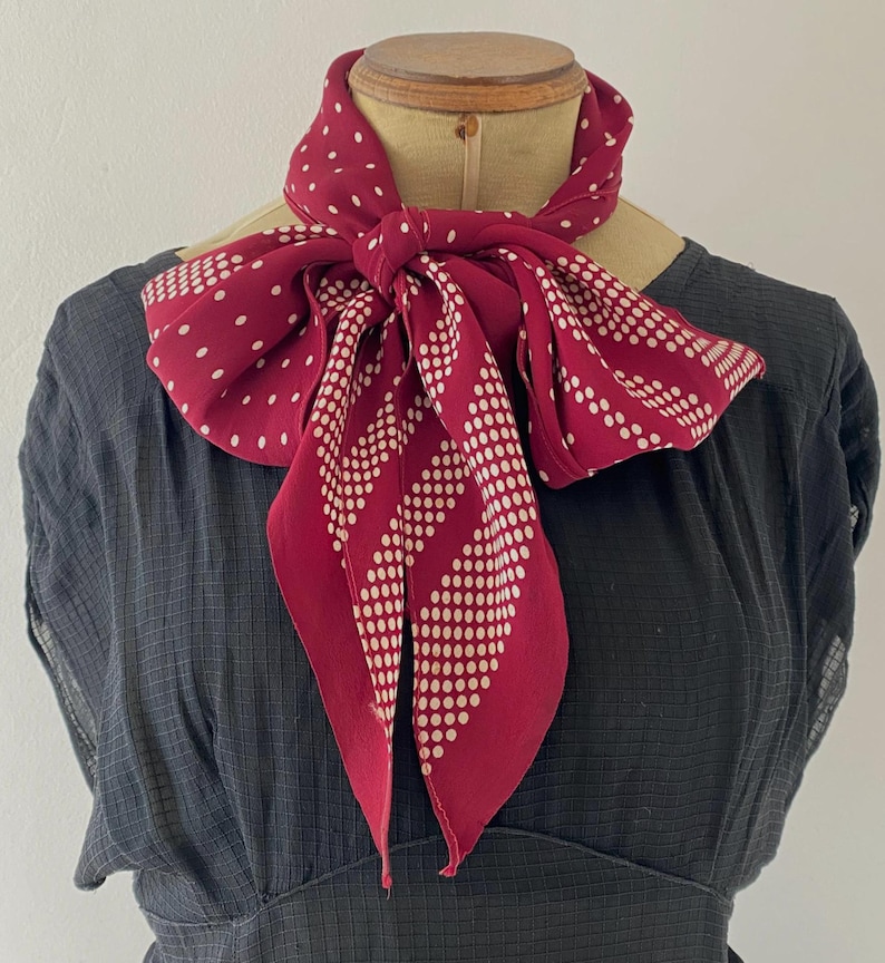 Puede incluir: Una bufanda burdeos con lunares blancos y un patr&oacute;n de rayas punteadas est&aacute; atada alrededor del cuello de un maniqu&iacute;. La bufanda est&aacute; hecha de una tela suave y fluida y est&aacute; anudada en un lazo. El fondo es un vestido gris oscuro.