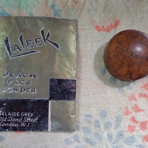 Könnte beinhalten: Ein Vintage-Puderdose aus braunem Material mit dem Wort "LALEK" auf der Oberseite. Die Dose liegt neben einem Papierbeutel mit den Worten "LALEK", "PEACH FACE POWDER" und "ADELAIDE GREY 27 Old Bond Street London. W.1."