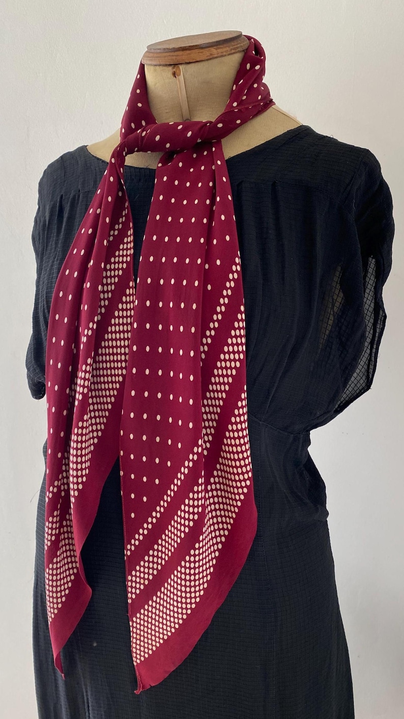 Puede incluir: Una bufanda burdeos con un estampado de lunares blancos y rayas diagonales de puntos blancos. La bufanda est&aacute; drapeada sobre una prenda negra, mostrando su dise&ntilde;o y contraste de color. La bufanda est&aacute; anudada al cuello.