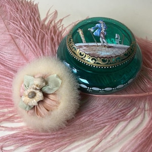 以下が含まれることがあります： 緑色のガラス製の小物入れとパフが特徴のヴィンテージバニティセット。小物入れには絵が描かれており、パフは丸く、ふわふわした外観と花の装飾が施されています。セットはピンクの羽の上に置かれています。