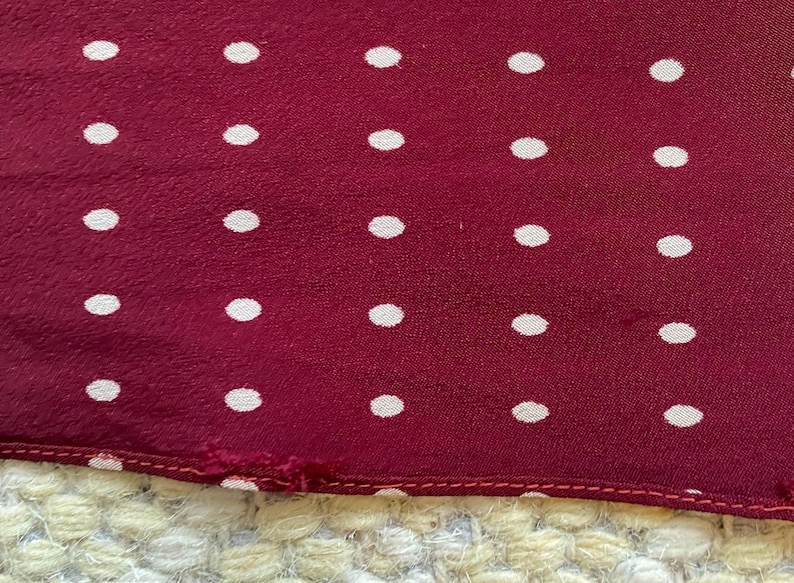 Puede incluir: Primer plano de una tela burdeos con un patr&oacute;n de lunares blancos espaciados uniformemente. La tela tiene un aspecto ligeramente texturizado y un borde cosido. Los lunares son peque&ntilde;os y circulares.