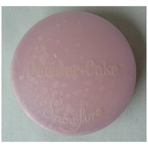 Può includere: Un compatto rotondo rosa con le parole "Powder-Cake" e "Snowfire" in rilievo sulla parte superiore.