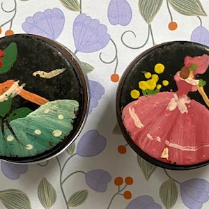 May include: Two circular vintage trinket boxes with hand-painted designs. One depicts a figure in a green dress, the other a figure in a pink dress. Both have black backgrounds and silver rims. Decorative and collectible items.