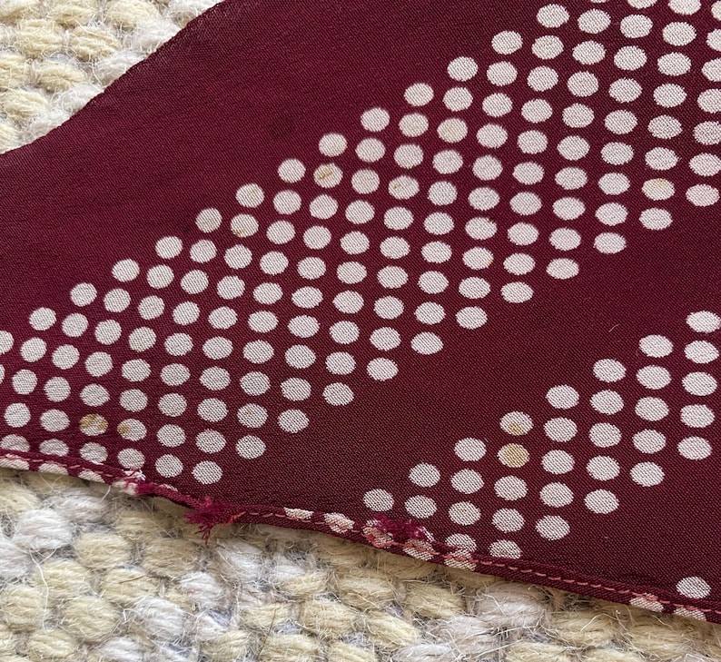 Puede incluir: Primer plano de una tela burdeos con un estampado de lunares blancos. La tela tiene un dise&ntilde;o diagonal de puntos blancos. El borde de la tela es visible, mostrando las costuras y un ligero deshilachado.