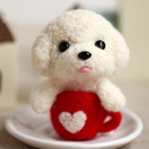 May include: A white felt dog holding a red felt mug with a white felt heart on it. The mug is sitting on a white saucer.