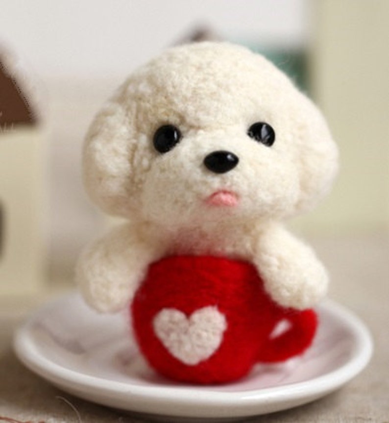 May include: A white felt dog holding a red felt mug with a white felt heart on it. The mug is sitting on a white saucer.