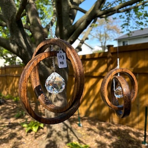 May include: Two wooden wind chimes with interlocking circular designs. Each chime features a faceted crystal pendant suspended from a string of beads. One chime has a tag with the numbers "103" and the letters "HD". The chimes are hanging from a tree.
