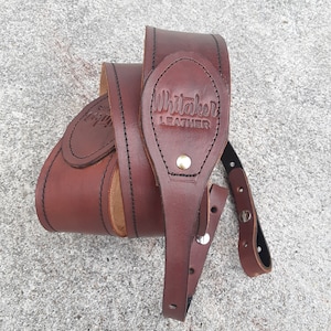 Puede incluir: Correa de guitarra de cuero marrón con un diseño cosido y un logotipo en relieve dorado que dice "Whitaker Leather".