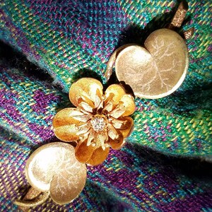 May include: A gold-toned brooch with a flower design and a small diamond in the center. The brooch is on a green and purple plaid fabric.