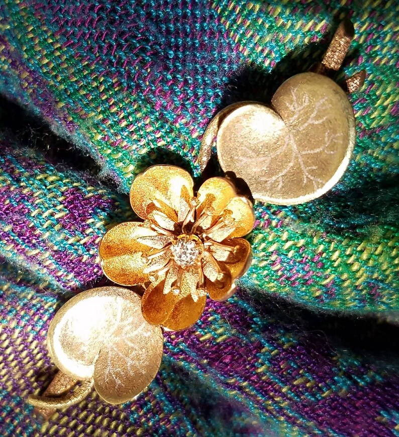 May include: A gold-toned brooch with a flower design and a small diamond in the center. The brooch is on a green and purple plaid fabric.
