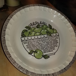 Könnte beinhalten: Ein weißer Keramik-Tortenteller mit schwarzem und weißem Rand. Der Teller zeigt eine grüne Apfelkuchen-Illustration mit dem Text "apple pie" und ein Rezept für Apfelkuchen.