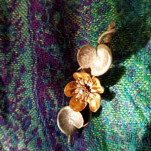 May include: A gold-toned pendant featuring a stylized flower with a small, clear gemstone in the center. The flower is attached to a stem with two round, gold-toned leaves.