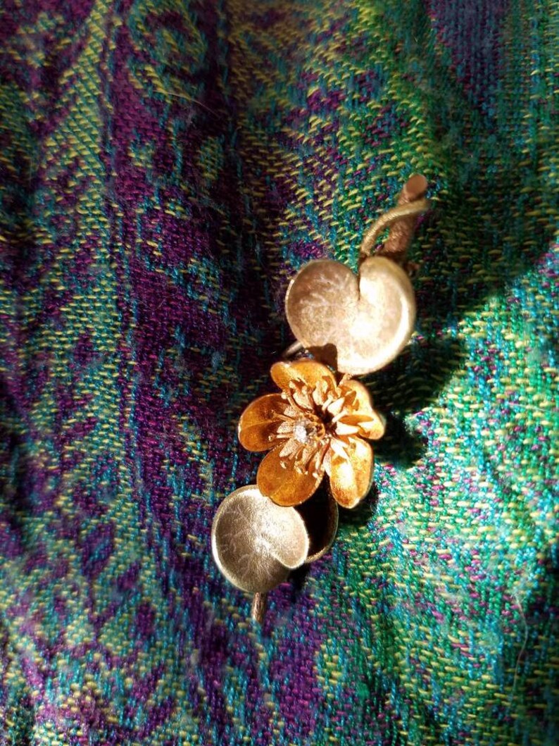 May include: A gold-toned pendant featuring a stylized flower with a small, clear gemstone in the center. The flower is attached to a stem with two round, gold-toned leaves.