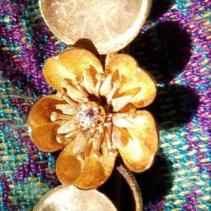 May include: A gold-toned brooch featuring a stylized flower with a single diamond in the center. The flower is surrounded by two round, leaf-shaped accents.