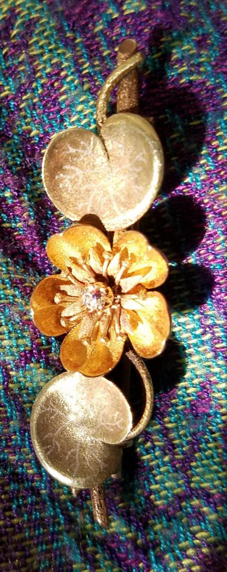 May include: A gold-toned brooch featuring a stylized flower with a single diamond in the center. The flower is surrounded by two round, leaf-shaped accents.
