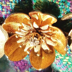 May include: A gold-colored flower brooch with a clear rhinestone center. The brooch is set against a purple and green plaid fabric.