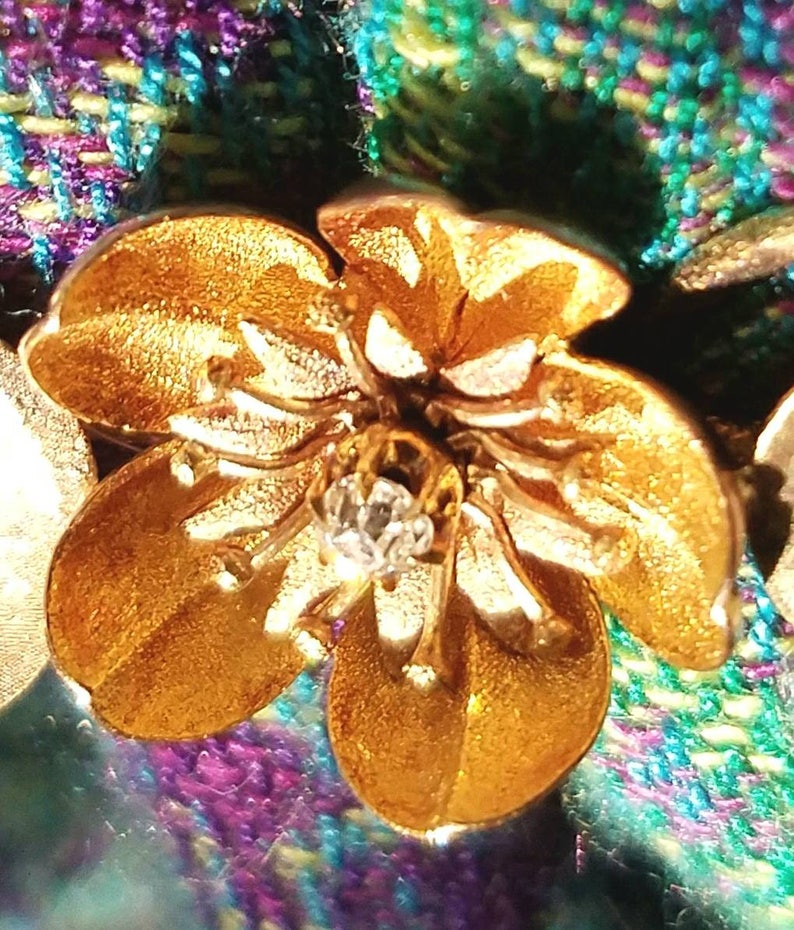 May include: A gold-colored flower brooch with a clear rhinestone center. The brooch is set against a purple and green plaid fabric.