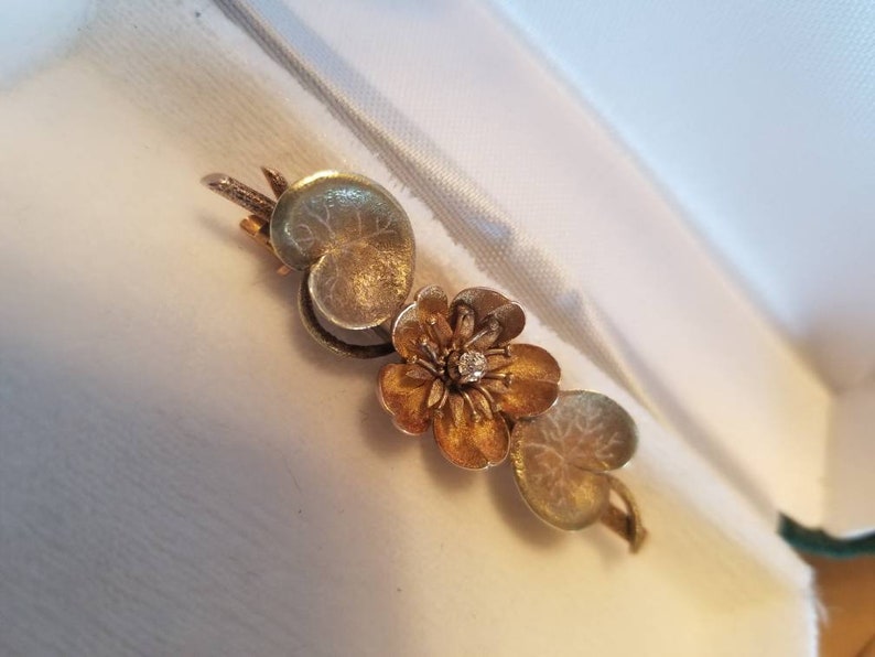 May include: A gold and silver brooch featuring a stylized flower with a single diamond in the center, surrounded by two leaves.