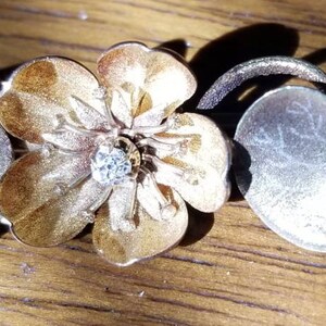 May include: A silver and gold floral jewelry piece with a flower and two leaves. The flower is a light gold color with a darker gold center. The leaves are silver and have a textured surface.