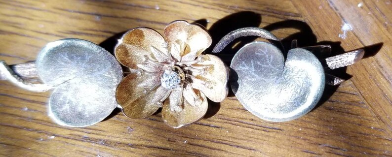 May include: A silver and gold floral jewelry piece with a flower and two leaves. The flower is a light gold color with a darker gold center. The leaves are silver and have a textured surface.