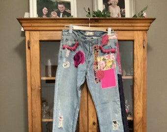 Op de afbeelding: Lichtblauwe denim jeans met patches en versieringen. De jeans hebben een distressed look met scheuren en gaten. De patches zijn gemaakt van verschillende stoffen en bevatten bloemenpatronen, roze pompoenen en een roze hart. De jeans hebben een rafelige zoom.