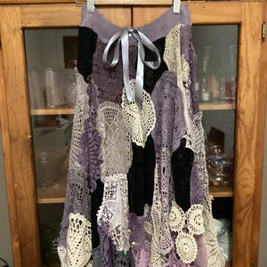 Könnte beinhalten: Ein langer, fließender Rock aus einem Patchwork aus gehäkelten Deckchen in den Farben Lila, Creme und Schwarz. Der Rock hat einen Gürtel an der Taille und ist perfekt für einen Boho- oder Vintage-Look.