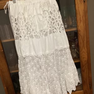 May include: A white skirt with detailed lace inserts. The skirt has a combination of solid fabric and openwork lace patterns, with a scalloped hem. The skirt is suspended from a wooden hanger.