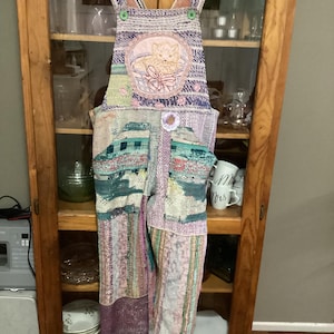 Può includere: Una salopette in patchwork con un gatto ricamato sul davanti. La salopette è realizzata con una varietà di tessuti in tonalità di viola, rosa, verde e blu.