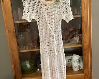 Könnte beinhalten: Ein cremefarbenes Häkelkleid mit offenem Gewebe und kurzen Ärmeln hängt an einem Holzbügel. Eine Perlenkette drapiert über dem Kleid. Zwei Stoffblumen sind auf der Schulter befestigt. Das Kleid wird vor einem Holzschrank ausgestellt.