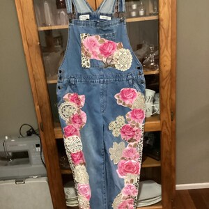 Può includere: Salopette di jeans blu con applicazioni floreali rosa e bianche e bordi in pizzo.