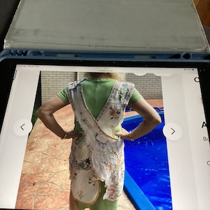 Könnte beinhalten: Eine weiße Häkel-Schürze mit Blumenstickerei und einem grünen Oberteil. Die Schürze hat eine Tasche und wird über einem grünen Shirt getragen. Die Person steht in der Nähe eines Schwimmbeckens.