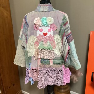 May include: A colorful patchwork jacket with a whimsical doll design on the back. The doll has a heart, flowers, and wings. The jacket is made of a variety of fabrics, including lace, knit, and woven materials.