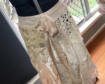 May include: A cream-colored skirt made of various lace and crochet panels, with a satin ribbon tie. The skirt is displayed on a black dress form. The skirt's design includes floral patterns and openwork details, creating a layered, textured look.