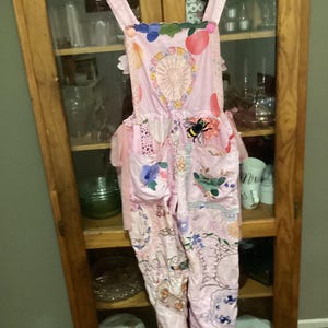 Puede incluir: Mono rosa con adornos florales y de abeja. El mono presenta un diseño de patchwork con encaje, bordados y apliques. Las correas son anchas y adornadas con acentos coloridos. El diseño general es caprichoso y femenino.