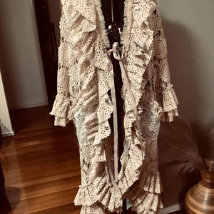 Puede incluir: Un cárdigan largo de crochet beige con un dobladillo y mangas con volantes. El cárdigan tiene cuello en V y se ata a la cintura con una cinta.