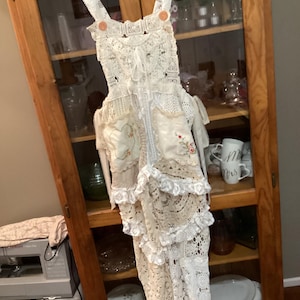 May include: A cream-coloured, handmade lace dress with a tiered skirt and adjustable straps. The dress features decorative floral embroidery on the pockets and is adorned with orange buttons. It hangs on a wooden hanger against a wooden cabinet.
