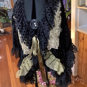 May include: A black velvet jacket with gold detailing and a large decorative button. The jacket has a polka dot pattern, ruffled trim, and gold ribbon. The garment is displayed on a black dress form.