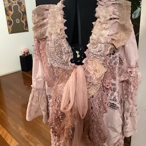 May include: A pink, layered jacket with intricate lace and fabric details. The jacket features a ruffled collar, floral embellishments, and a flowing, sheer bow. The garment is displayed on a black dress form, showcasing its unique design.