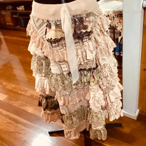 May include: A multi-layered skirt in pink, cream, and olive green. The skirt is made from lace and patterned fabrics, with a cream-colored tie. It is displayed on a black mannequin stand. The skirt has a vintage aesthetic.