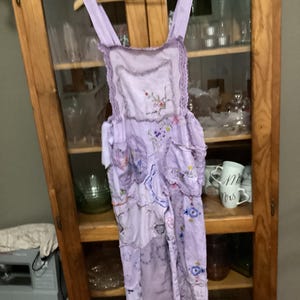 Puede incluir: Un delantal lavanda con bordados florales y ribetes de encaje cuelga de una percha de madera. El delantal presenta múltiples capas de tela y bolsillos, con una estética delicada y vintage. El delantal se exhibe frente a un armario de madera.