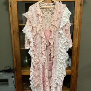 以下が含まれることがあります： フリルが付いた長いピンクと白のレースのローブ。ローブは木製のハンガーにかけられ、木製のキャビネットの前にある。