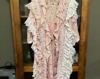 May include: A long, pink and white lace robe with ruffles. The robe is hanging on a wooden hanger in front of a wooden cabinet.