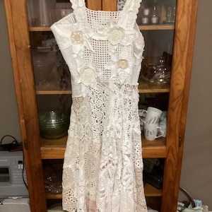 Peut inclure: Une robe blanche en crochet avec des embellissements floraux. La robe a un corsage ajusté et une jupe ample.