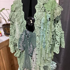 以下が含まれることがあります： 重ね着のデザインが施された緑色のクロシェカーディガン。カーディガンはさまざまな種類のレースで作られており、流れるようなボヘミアンスタイルです。