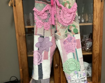 Op de afbeelding: Een broek van patchwork met roze gehaakte accenten en een roze strik. De broek is gemaakt van verschillende stoffen, waaronder denim, katoen en kant.