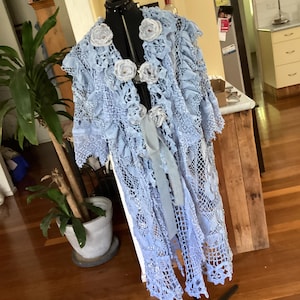以下が含まれることがあります： ライトブルーのオープンフロントかぎ針編みダスター、花のアクセント付き。 長く流れるような衣服は、黒いマネキンに展示されています。 ダスターは繊細なレース模様で、白い布の花で飾られています。