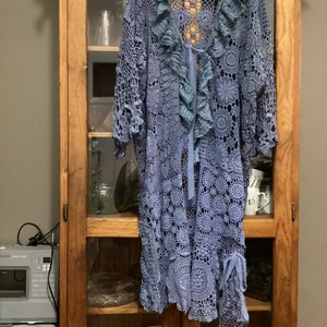 May include: A pale blue, open-front crochet garment with a floral design and frilled trim. The garment is hanging on a wooden hanger inside a wooden cabinet. The sleeves and front have decorative ties.