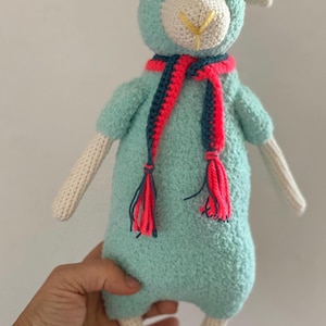 May include: A turquoise sheep doll with a white face, ears, and limbs. The sheep is wearing a scarf with red, blue, and pink tassels. The doll is made of a soft, textured material.