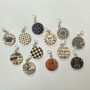 May include: Twelve round, wooden charms with different patterns and designs. The charms have silver metal rings attached to them. The patterns include a clock face, polka dots, a checkerboard, a crown, a number grid, and a floral design.